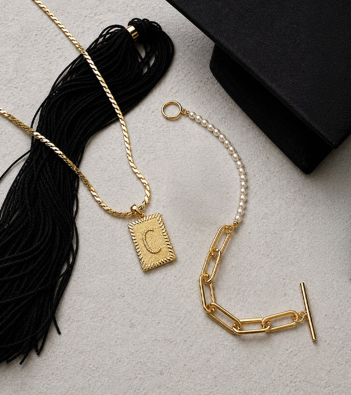 Gold necklace with a letter 'C' pendant and black tassel on a light gray surface.